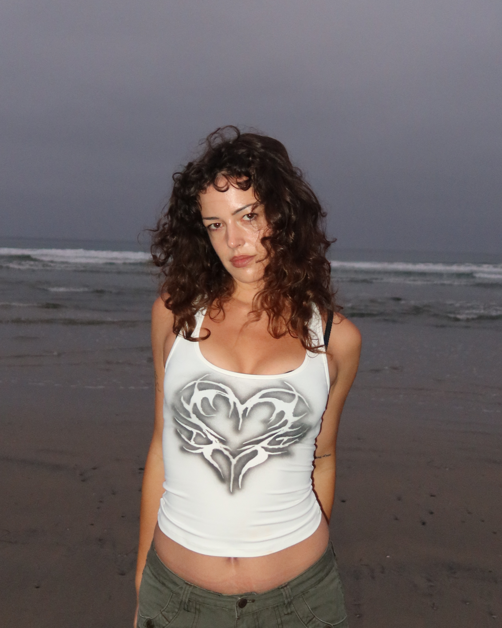 A woman with curly brown hair stands on a beach wearing a white cropped tank top with a gray winged heart graphic and olive green cargo pants, set against a dark, overcast sky and ocean.