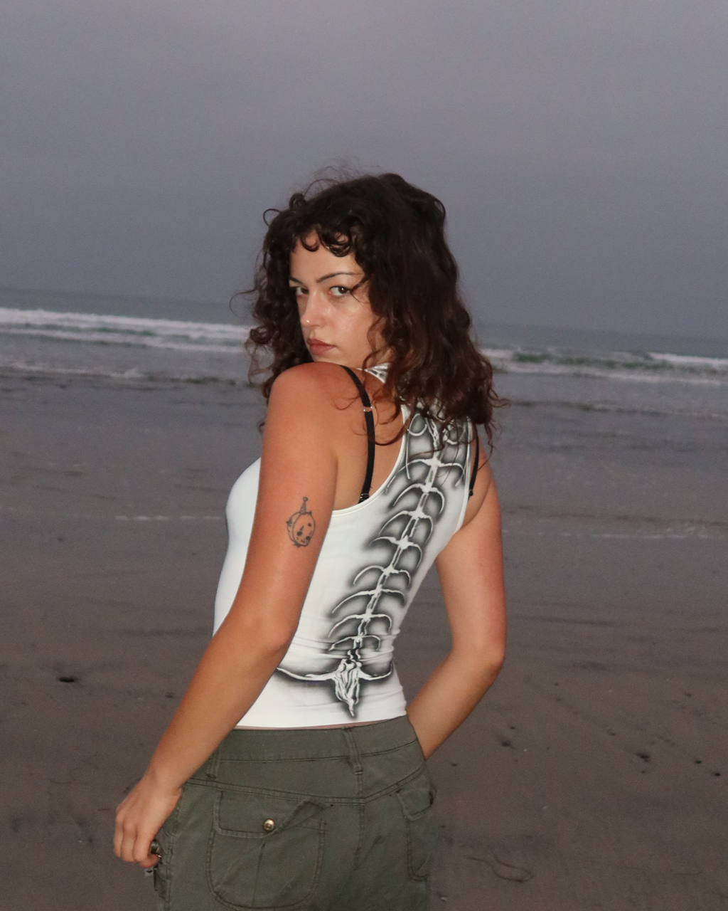 A woman stands on a sandy beach facing away from the camera, wearing a white tank top with a black graphic spine print and olive green cargo pants under an overcast sky.