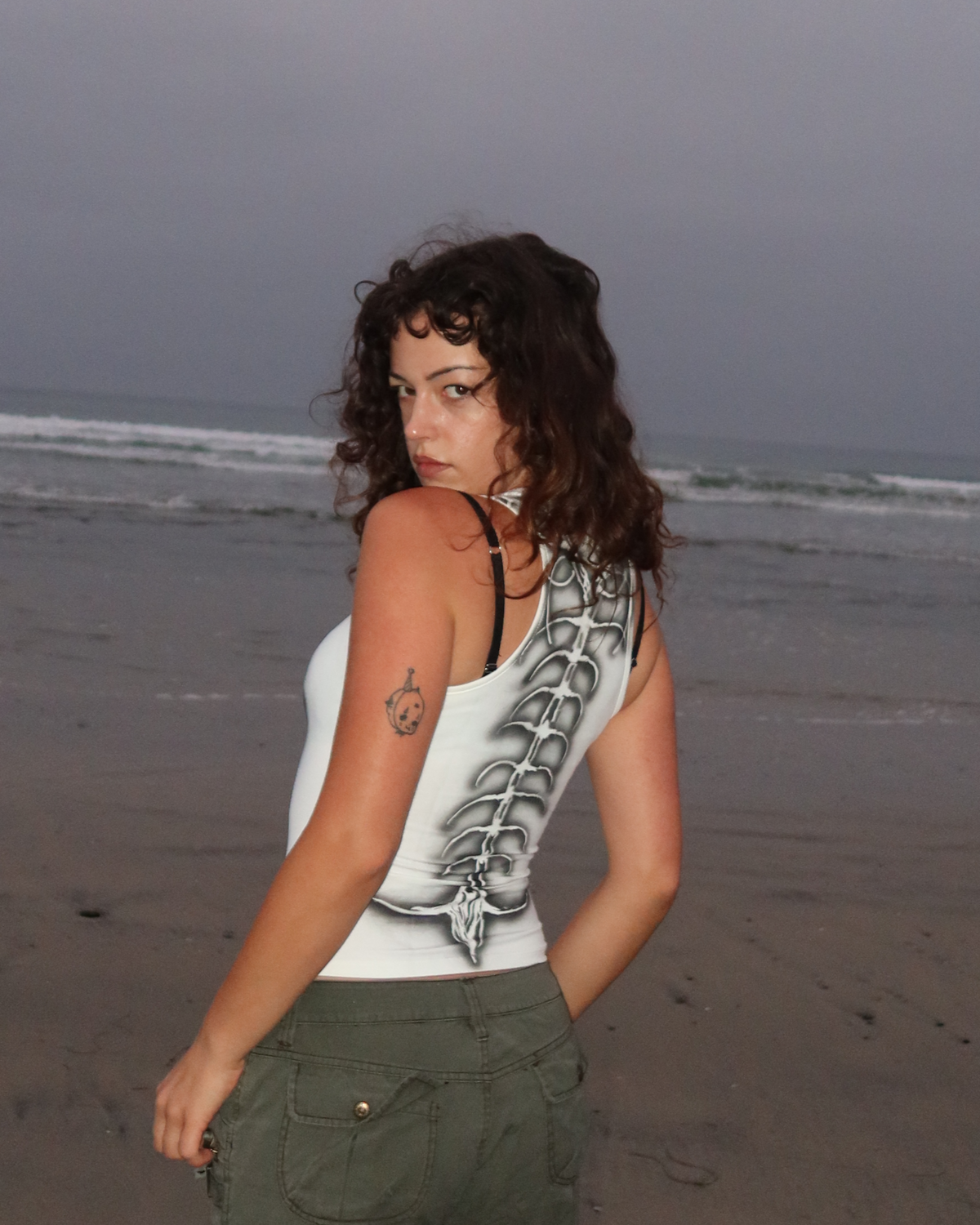 A woman stands on a sandy beach facing away from the camera, wearing a white tank top with a black graphic spine print and olive green cargo pants under an overcast sky.