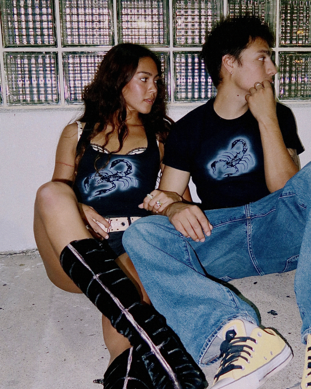A couple sitting together in front of a glass block wall. The woman wears a black tank top with a blue graphic, black thigh-high boots, and a silver belt.