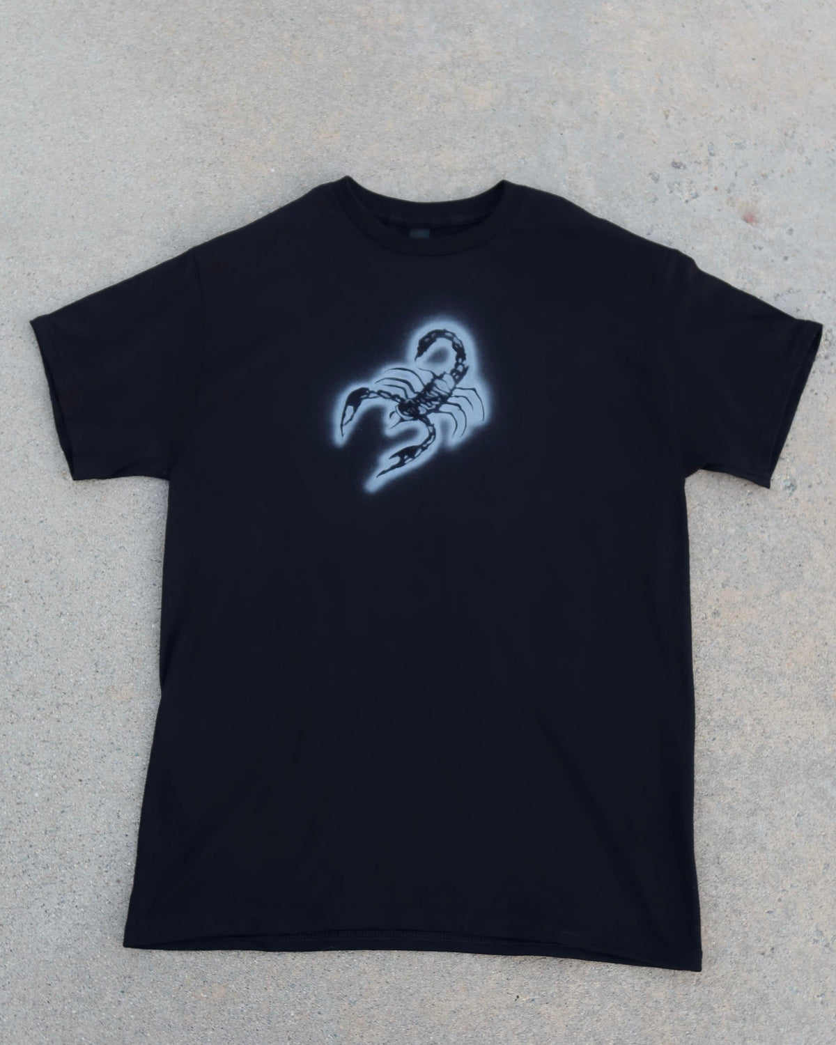 A black short-sleeve t-shirt with a glowing blue scorpion graphic on the chest, laid flat on a textured gray concrete surface.